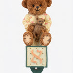 Teddy bear with a smaller teddy bear on its lap, sitting on a mezuzah case with a pink bow on a white background.