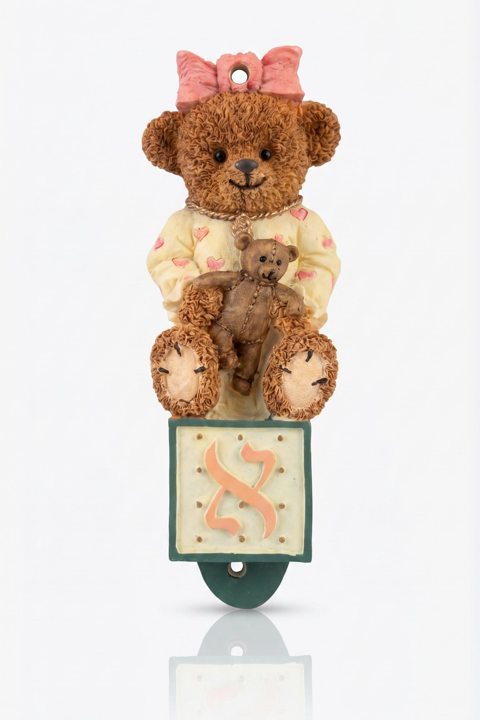 Teddy bear with a smaller teddy bear on its lap, sitting on a mezuzah case with a pink bow on a white background.