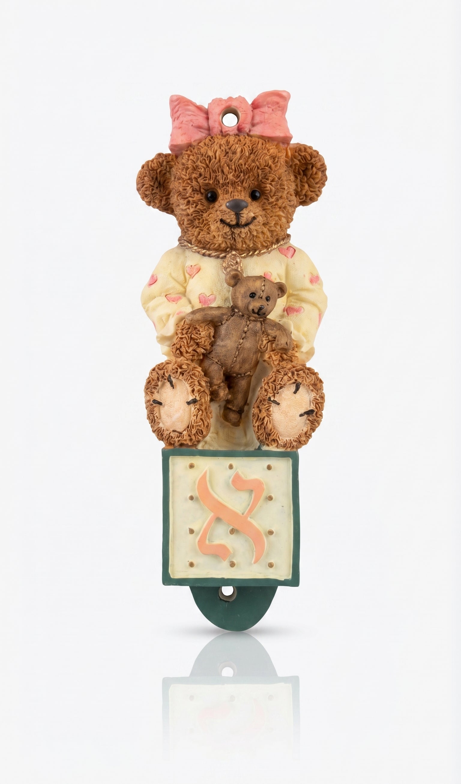 Teddy bear with a smaller teddy bear on its lap, sitting on a mezuzah case with a pink bow on a white background.