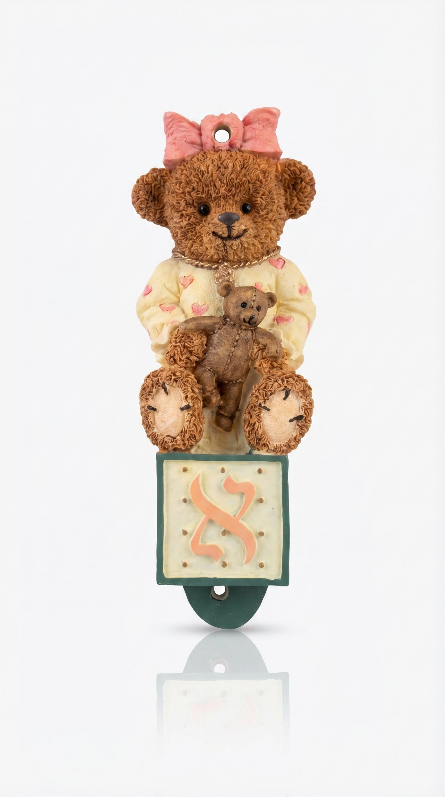 Teddy bear with a smaller teddy bear on its lap, sitting on a mezuzah case with a pink bow on a white background.