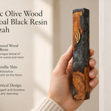 Hand holding a 7.5-inch olive wood and charcoal resin mezuzah case with gold Shin detail against a soft neutral background.