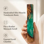 Hand holding an olive wood mezuzah case featuring flowing emerald green resin and a gold Shin symbol in a premium editorial setting.