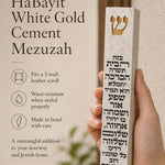 Hand holding a Birkat HaBayit White Gold Cement Mezuzah case with a raised gold Shin and full Hebrew home-blessing text printed vertically across the slim white cement body, shown in a warm neutral home setting