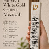 Hand holding a Birkat HaBayit White Gold Cement Mezuzah case with a raised gold Shin and full Hebrew home-blessing text printed vertically across the slim white cement body, shown in a warm neutral home setting