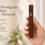 A person holds a brown woodgrain-look acrylic mezuzah with a silver crown and Shin, presented in an editorial lifestyle style.
