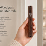 A hand holds a classic woodgrain acrylic mezuzah case featuring a silver Hebrew Shin on a bright, minimalist background.