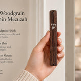 A hand holds a classic woodgrain acrylic mezuzah case featuring a silver Hebrew Shin on a bright, minimalist background.