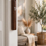 Classic woodgrain acrylic mezuzah with a silver Shin mounted on a white doorframe beside a warm living room with a potted olive tree, showing the dark wood-look finish and slim profile of this waterproof Israeli mezuzah case in place