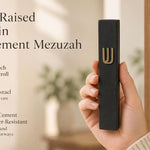 Close up of a hand holding a six-inch black cement mezuzah case with a prominent gold Hebrew Shin character