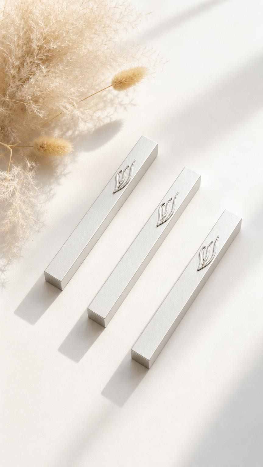 Three Eternal Flame silver aluminum mezuzah cases arranged on a warm neutral surface with dried florals, featuring raised silver ner tamid and stylized Shin emblems