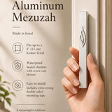 Eternal Flame silver aluminum mezuzah case held against a light-colored doorpost, showing its slim profile and raised ner tamid emblem; made in Israel, waterproof, and sized for up to a 5-inch kosher scroll