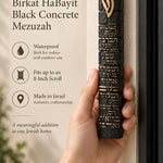Hand holding an extra large textured black concrete mezuzah with gold Birkat HaBayit lettering and a raised gold Shin against the doorway of a home