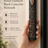 Hand holding an extra large textured black concrete mezuzah with gold Birkat HaBayit lettering and a raised gold Shin against the doorway of a home