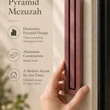 Extra Large Modern Burgundy Pyramid Mezuzah held beside a doorpost, with the muted burgundy wine-red aluminum mezuzah cover shown close enough to reveal its three stepped bars and rounded lower edge alongside product-name and design callouts for this minimalist Judaica piece