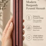 A hand presents the Extra Large Modern Burgundy Pyramid Mezuzah with its muted burgundy wine-red aluminum mezuzah cover facing forward, highlighting the clean graduated bars and geometric vertical profile beside design illustrations for a minimalist Jewish doorway accent