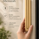 Extra Large Modern Gold Pyramid Mezuzah held beside a doorpost, with the warm metallic gold aluminum mezuzah cover shown close enough to reveal its three stepped bars and rounded lower edge alongside product-name and design callouts for this minimalist Judaica piece