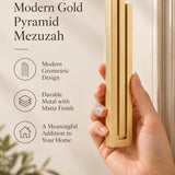 A hand presents the Extra Large Modern Gold Pyramid Mezuzah with its warm metallic gold aluminum mezuzah cover facing forward, highlighting the clean graduated bars and geometric vertical profile beside design illustrations for a minimalist Jewish doorway accent