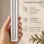 A hand presents the Extra Large Modern Silver Pyramid Mezuzah with its softly brushed silver aluminum mezuzah cover facing forward, highlighting the clean graduated bars and geometric vertical profile beside design illustrations for a minimalist Jewish doorway accent