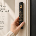 Person holding an 8.5-inch black concrete mezuzah with gold Birkat HaBayit lettering and a raised gold Shin in a hand against the entrance doorpost of a home