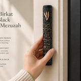 Person holding an 8.5-inch black concrete mezuzah with gold Birkat HaBayit lettering and a raised gold Shin in a hand against the entrance doorpost of a home