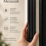 Giant Modern Black Pyramid Mezuzah held beside a doorpost, with the deep matte black aluminum mezuzah cover shown close enough to reveal its three stepped bars and rounded lower edge alongside product-name and design callouts for this architectural Judaica piece