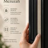 Giant Modern Black Pyramid Mezuzah held beside a doorpost, with the deep matte black aluminum mezuzah cover shown close enough to reveal its three stepped bars and rounded lower edge alongside product-name and design callouts for this architectural Judaica piece