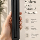 Hand holding the Giant Modern Black Pyramid Mezuzah upright, revealing its deep matte black aluminum mezuzah cover, slender three-bar Pyramid design and graduated top beside illustrated product features for architectural Jewish home Judaica