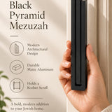 A hand presents the Giant Modern Black Pyramid Mezuzah with its deep matte black aluminum mezuzah cover facing forward, highlighting the clean graduated bars and geometric vertical profile beside design illustrations for a architectural Jewish doorway accent