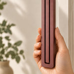 A hand displays the Giant Modern Burgundy Pyramid Mezuzah beside a doorway, showing the rich burgundy finish of this aluminum mezuzah cover, its long graduated bars and rounded lower edge in a architectural expression of Jewish home Judaica