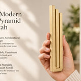 Giant Modern Gold Pyramid Mezuzah pictured in hand as a warm metallic gold aluminum mezuzah cover with three graduated bars and a architectural stepped design, alongside its name and illustrated product features in a wide social-sharing image
