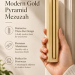 A hand presents the Giant Modern Gold Pyramid Mezuzah with its warm metallic gold aluminum mezuzah cover facing forward, highlighting the clean graduated bars and geometric vertical profile beside design illustrations for a architectural Jewish doorway accent
