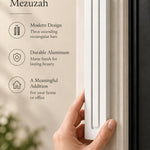 Giant Modern White Pyramid Mezuzah held beside a doorpost, with the smooth matte white aluminum mezuzah cover shown close enough to reveal its three stepped bars and rounded lower edge alongside product-name and design callouts for this architectural Judaica piece