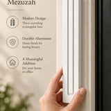 Giant Modern White Pyramid Mezuzah held beside a doorpost, with the smooth matte white aluminum mezuzah cover shown close enough to reveal its three stepped bars and rounded lower edge alongside product-name and design callouts for this architectural Judaica piece