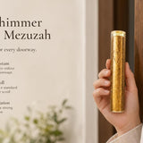 Gold Shimmer acrylic mezuzah case held beside a modern doorway, featuring a clear cylindrical acrylic body filled with sparkling gold accents and a subtle Hebrew Shin design. Made in Israel, this contemporary gold mezuzah case fits a 5-inch kosher mezuzah scroll and works beautifully on indoor or protected exterior doorways