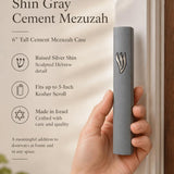 Handheld gray cement mezuzah case with a raised sculptural silver-tone Shin, shown beside product details to illustrate the slim proportions, smooth cement finish, minimalist design, and modern Jewish doorway style