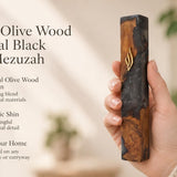 Person holding a handcrafted olive wood and black resin mezuzah featuring a gold Shin letter in a warm, naturally lit setting.