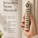 Close-up of person holding Jerusalem Stone Mezuzah, showing detailed black Hebrew lettering and cylindrical shape, on a natural background.