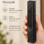 Close-up of a hand holding an embossed black concrete mezuzah case with gold-tone Shin detail against a neutral background.