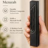 Close-up of a hand holding an embossed black concrete mezuzah case with gold-tone Shin detail against a neutral background.