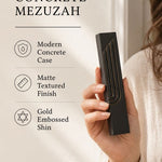 Close-up of a person holding a minimalist black concrete mezuzah with a sculpted gold-edged Shin symbol in a soft light.