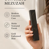 Close-up of a person holding a minimalist black concrete mezuzah with a sculpted gold-edged Shin symbol in a soft light.