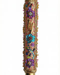 Intricate handmade Mezuzah decorated with colorful sequins and stones, mounted on a clean white background with soft reflection.