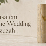 Jerusalem Stone Wedding mezuzah case displayed against a natural stone background, featuring warm cream Jerusalem stone, a flowing carved center line, Hebrew lettering along the side, and a gold Hebrew Shin on the front. A meaningful Jewish wedding, housewarming, or new home mezuzah gift with a timeless Jerusalem-inspired design