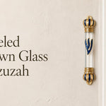 Jeweled Crown glass mezuzah case mounted beside a doorway, featuring a clear glass center, royal blue and gold crown-shaped end caps, sparkling crystal accents, and a large blue Hebrew Shin outlined in gold. This ornate glass Jewish mezuzah case creates an elegant statement for a home entrance or interior doorway