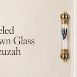 Jeweled Crown glass mezuzah case mounted beside a doorway, featuring a clear glass center, royal blue and gold crown-shaped end caps, sparkling crystal accents, and a large blue Hebrew Shin outlined in gold. This ornate glass Jewish mezuzah case creates an elegant statement for a home entrance or interior doorway