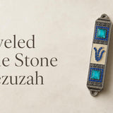 Jeweled Blue Stone pewter mezuzah case mounted on a light wall, featuring an antique silver-tone finish, bright turquoise square stones, rows of blue crystals, ornate detailing, and a jeweled Hebrew Shin. This decorative blue Jewish mezuzah case adds rich color and traditional Judaica style to a home doorway