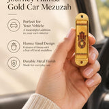 Hand holding the Journey Hamsa Gold Car Mezuzah mezuzah cover inside a vehicle, showing a brown Hamsa hand with a Star of David in its palm, warm gold-colored panels and patterned borders in a rounded stainless-steel frame beside the product name and illustrated features for this decorative Jewish travel Judaica piece