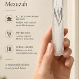 Kotel Stonework Clear Silver Shin Mezuzah mezuzah cover held in one hand beside its product name and illustrated features, with a clear plastic body, molded stone-block texture and an elongated silver-colored Hebrew Shin above the rounded bottom closure clearly visible in a close view of the molded texture and Hebrew design