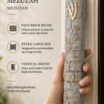 A hand holds an extra large gray concrete mezuzah featuring gold Hebrew lettering and Birkat HaBayit text in a bright, airy setting.
