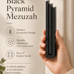 Hand holding the Large Modern Black Pyramid Mezuzah upright, revealing its deep matte black aluminum mezuzah cover, slender three-bar Pyramid design and graduated top beside illustrated product features for contemporary Jewish home Judaica